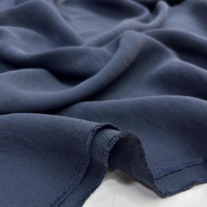 May include: A piece of dark slate blue fabric with a matte finish and a visible woven texture. The material is draped in soft, flowing folds, highlighting the depth of the deep blue color and the slightly frayed, raw edges of the textile.