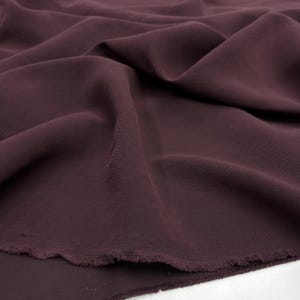 May include: A solid dark plum colored fabric with a smooth, matte texture. The material is draped in soft, flowing folds, creating deep shadows and highlights across the surface. The bottom edge shows a raw, unfinished frayed hemline.