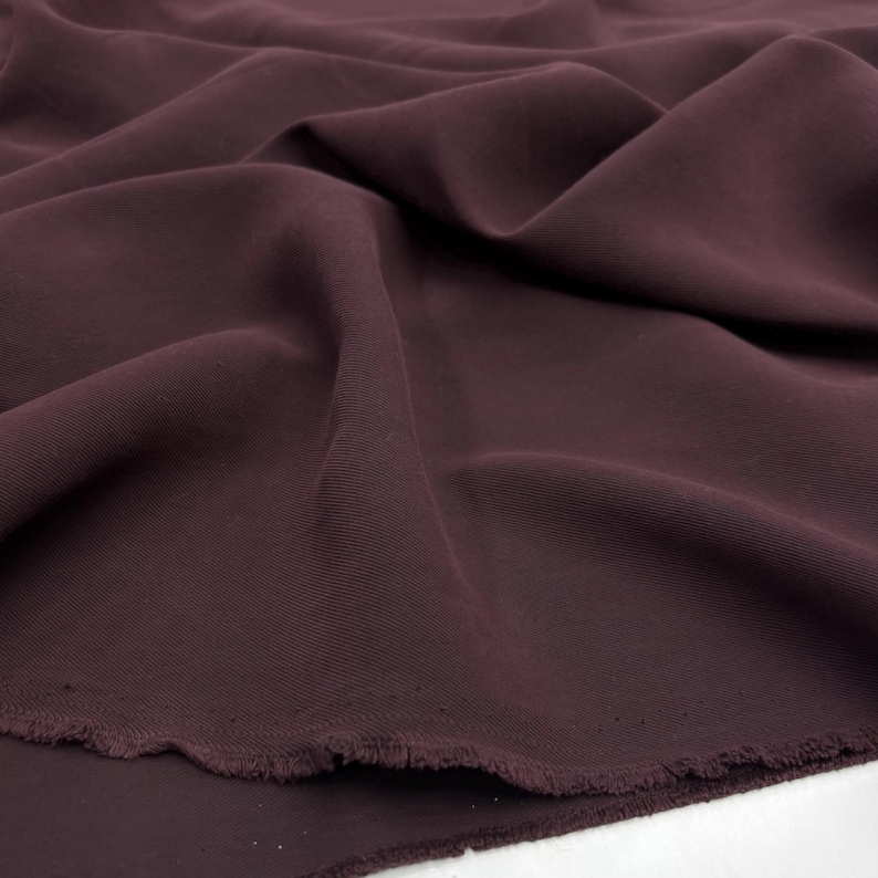 May include: A solid dark plum colored fabric with a smooth, matte texture. The material is draped in soft, flowing folds, creating deep shadows and highlights across the surface. The bottom edge shows a raw, unfinished frayed hemline.
