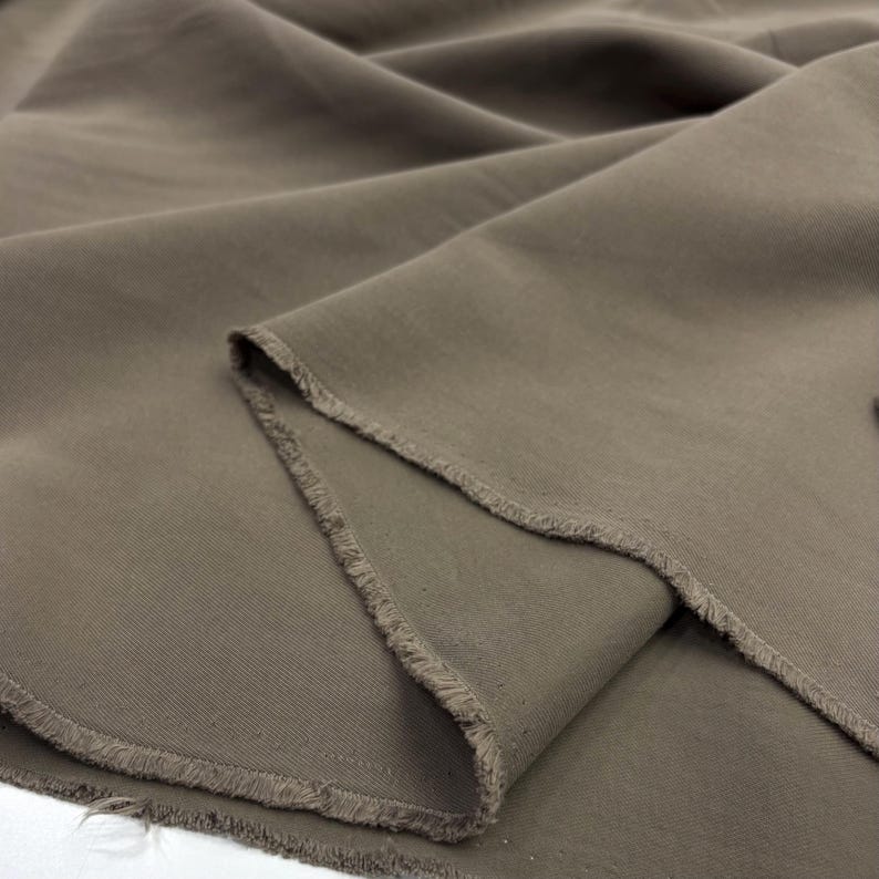 May include: A length of solid taupe-colored fabric with a visible weave and a raw, frayed edge. The material is draped in soft folds, highlighting the matte texture and the consistent earthy brown tone across the entire surface of the textile.