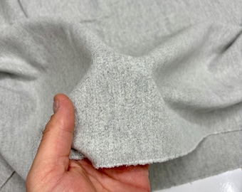 May include: Close-up of a light gray fabric, possibly cotton or a similar material. The fabric is held in a hand, showing its texture and the raw edge of the cut. The material appears soft and slightly textured.