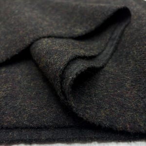 May include: Close-up of a dark grey fabric with a textured, woven appearance. The fabric has a slightly mottled effect with hints of brown and green fibres, creating a heathered look. The material is folded, revealing its thickness and drape.