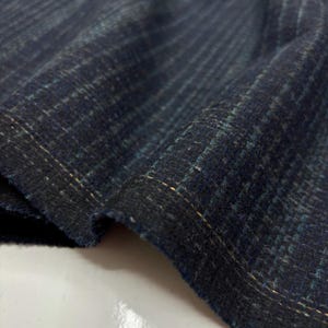 Puede incluir: Primer plano de una tela azul oscuro con un sutil patrón de cuadros. La tela tiene un aspecto texturizado con hilos visibles en tonos de azul, turquesa y dorado. Los bordes de la tela son visibles, mostrando el tejido y la textura.