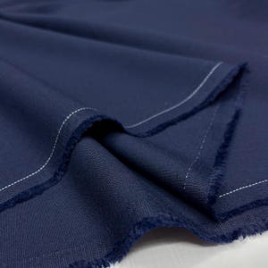 Könnte beinhalten: Nahaufnahme eines gefalteten, marineblauen Stoffes. Der Stoff hat eine sichtbare Textur und Webart. Weiße Nähte sind entlang der ausgefransten Kanten zu sehen. Geeignet für Kleidung oder Polsterung.