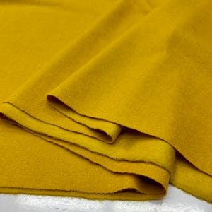 Puede incluir: Primer plano de una tela amarilla mostaza doblada. La tela tiene un aspecto suave y texturizado con pliegues y capas visibles. Los bordes de la tela son visibles, mostrando el grosor y la construcción del material.