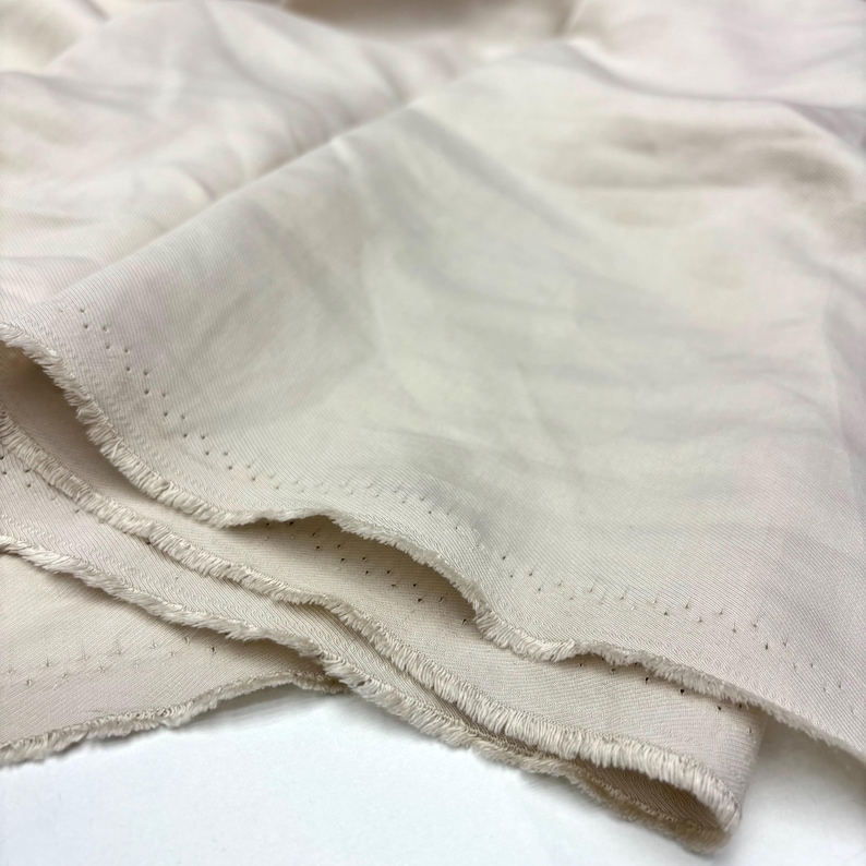 May include: A length of off-white fabric with a smooth, matte texture and visible folds. The edges of the material are frayed and feature a distinct row of small, evenly spaced pinhole perforations running along the selvage of the textile.
