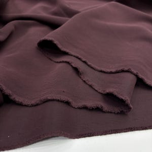 May include: A length of deep plum-colored fabric with a smooth, matte texture. The material is draped in soft, flowing folds, revealing a raw, unfinished edge with visible frayed threads along the cut side of the textile piece.