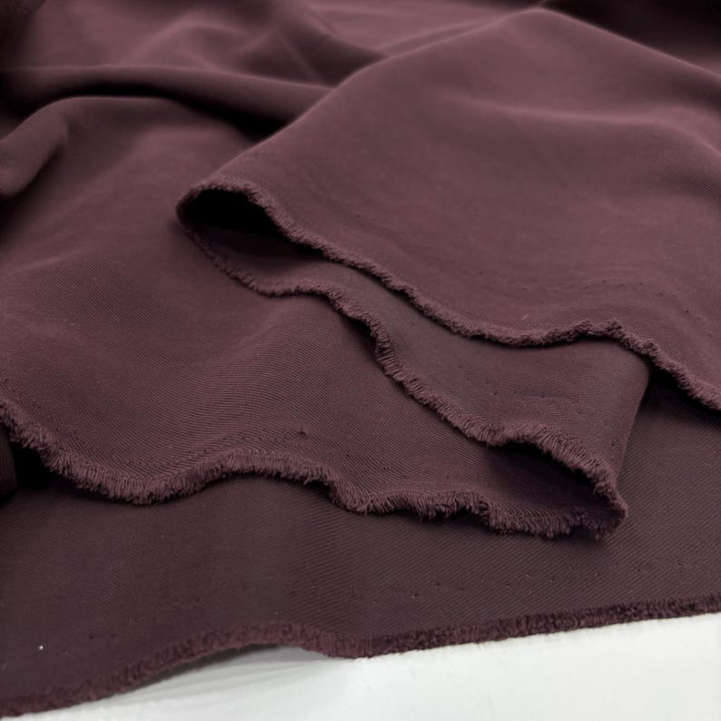 May include: A length of deep plum-colored fabric with a smooth, matte texture. The material is draped in soft, flowing folds, revealing a raw, unfinished edge with visible frayed threads along the cut side of the textile piece.
