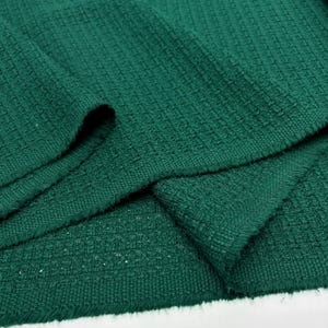 May include: Close-up of a folded piece of dark green fabric. The fabric has a textured weave pattern, with visible threads creating a slightly uneven surface. The folds create shadows, highlighting the texture and depth of the material.