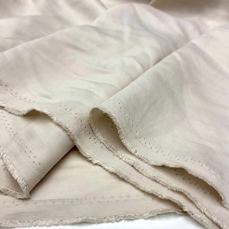 May include: A length of off-white, cream-colored fabric with a smooth, slightly lustrous surface. The material features a visible twill weave pattern and raw, frayed edges along the sides, which include a series of small, evenly spaced pinholes.