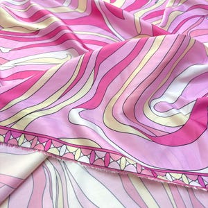 May include: Pink fabric featuring a swirling, abstract design in shades of pink, white, and yellow. The fabric has a decorative border with a repeating geometric pattern. The material is likely silk or a similar fabric.