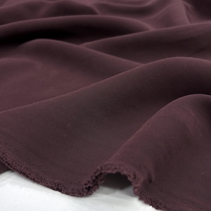 May include: A deep plum-colored fabric with a visible twill weave texture. The material is draped in soft, flowing folds, showcasing the matte finish and the slightly frayed, raw edges along the bottom border of the textile piece.