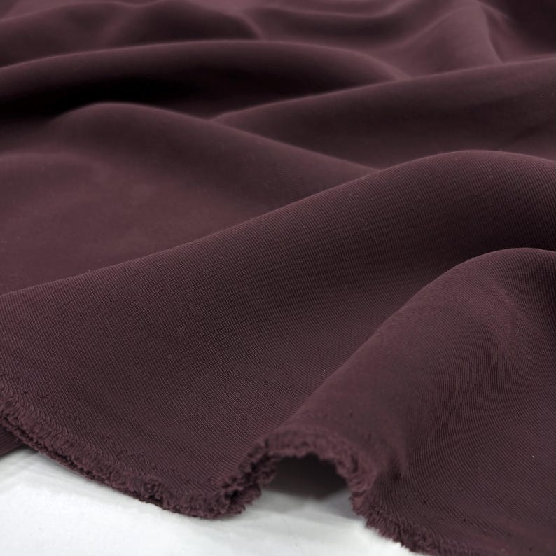 May include: A deep plum-colored fabric with a visible twill weave texture. The material is draped in soft, flowing folds, showcasing the matte finish and the slightly frayed, raw edges along the bottom border of the textile piece.