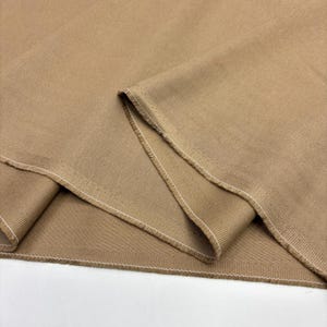 Puede incluir: Una tela de color tostado sólido con una textura tejida visible y un patrón de sarga distintivo. El material está doblado para revelar los bordes crudos y sin rematar a lo largo de las costuras, reforzados con una puntada overlock blanca.