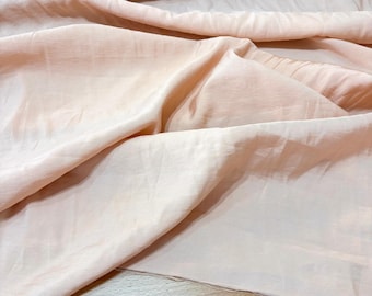 May include: Close-up of a roll of peach-colored fabric with a soft, flowing texture. The fabric is draped and folded, creating shadows and highlights. The material appears lightweight and possibly suitable for clothing or home decor.