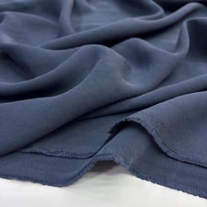 May include: A length of solid slate blue fabric with a soft, matte finish and a visible twill weave texture. The material is draped in loose, flowing folds, showcasing the flexible nature of the textile and its raw, unfinished frayed edges.