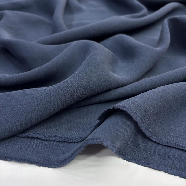 May include: A length of solid slate blue fabric with a soft, matte finish and a visible twill weave texture. The material is draped in loose, flowing folds, showcasing the flexible nature of the textile and its raw, unfinished frayed edges.
