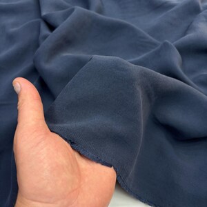 May include: A piece of solid dark blue fabric with a matte finish and a soft, draped texture. The material is shown in folds, highlighting the deep navy blue color and the smooth, consistent surface of the textile. The edges appear slightly frayed.
