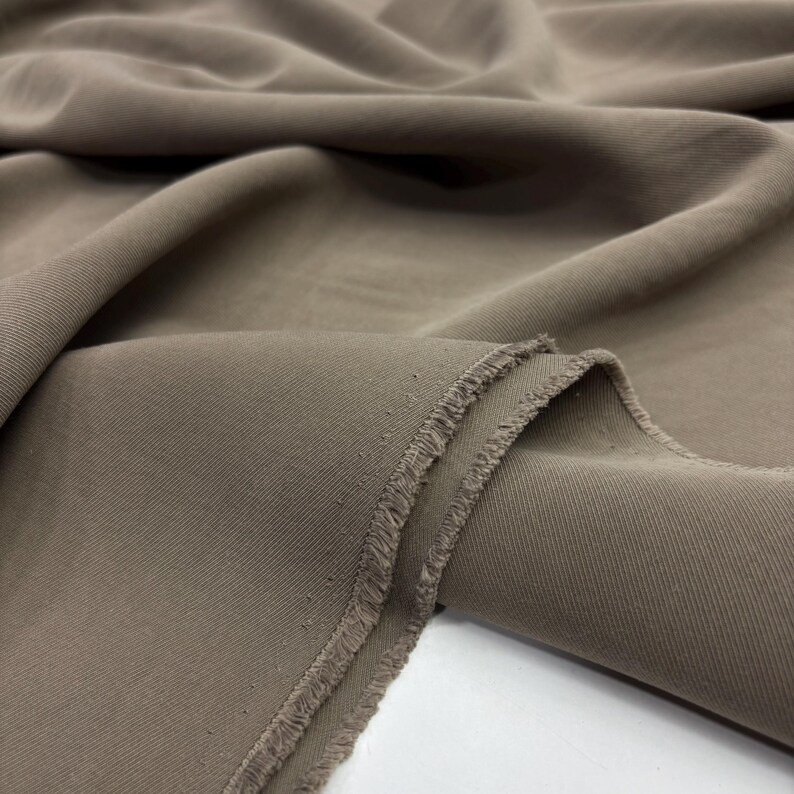 May include: A length of solid taupe-colored fabric featuring a distinct diagonal twill weave pattern. The material is draped in soft folds, revealing a raw, frayed edge along the cut side of the textile, showcasing the texture and weave.