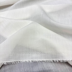 May include: Close-up of white fabric with a textured weave. The fabric is folded, creating shadows and highlights. The edge of the fabric is frayed, and there are small holes along the edge. The fabric appears to be lightweight, possibly linen or cotton.
