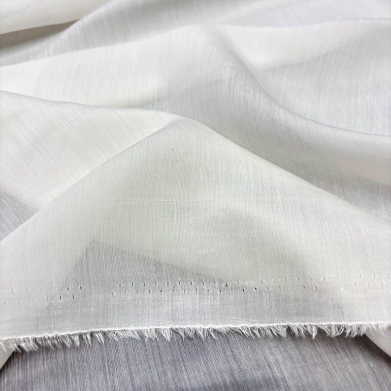 May include: Close-up of white fabric with a textured weave. The fabric is folded, creating shadows and highlights. The edge of the fabric is frayed, and there are small holes along the edge. The fabric appears to be lightweight, possibly linen or cotton.