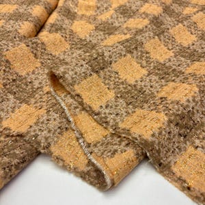 May include: A textured fabric featuring a checkered pattern in shades of light orange and brown. The material has a woven appearance with metallic gold threads interspersed throughout the weave, creating a subtle shimmer across the surface.