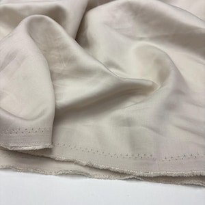 May include: A length of off-white fabric with a smooth, lustrous finish and a visible diagonal twill weave pattern. The material is draped in soft folds, revealing a raw, frayed edge along the bottom with a series of small, evenly spaced holes.