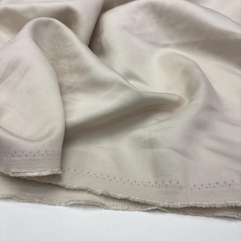 May include: A length of off-white fabric with a smooth, lustrous finish and a visible diagonal twill weave pattern. The material is draped in soft folds, revealing a raw, frayed edge along the bottom with a series of small, evenly spaced holes.