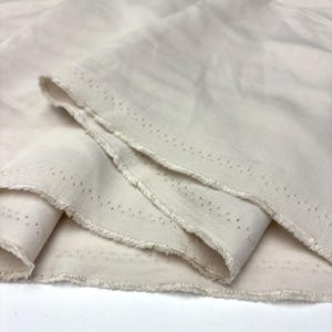 May include: A length of off-white, cream-colored fabric with a visible twill weave texture. The material features a raw, frayed edge along the selvage, which is marked by a series of small, evenly spaced dark pinhole dots running along the border.