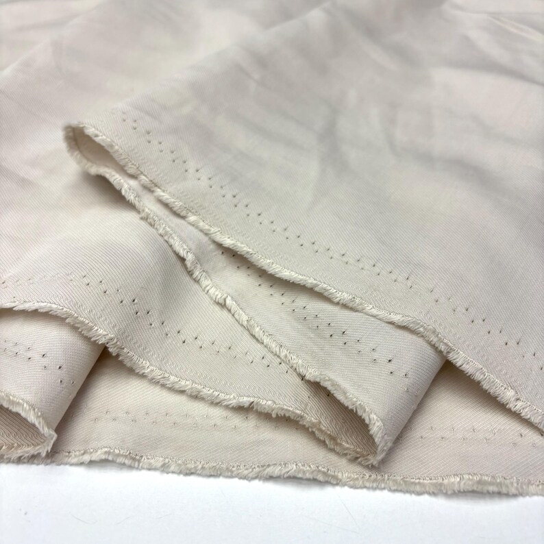 May include: A length of off-white, cream-colored fabric with a visible twill weave texture. The material features a raw, frayed edge along the selvage, which is marked by a series of small, evenly spaced dark pinhole dots running along the border.