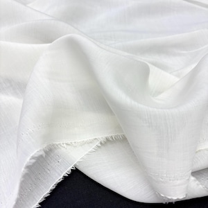 May include: Close-up of white fabric with a soft, flowing texture. The material is draped, showing folds and subtle variations in light and shadow. The edges of the fabric are unfinished, with a frayed appearance. The fabric appears to be lightweight and semi-sheer.