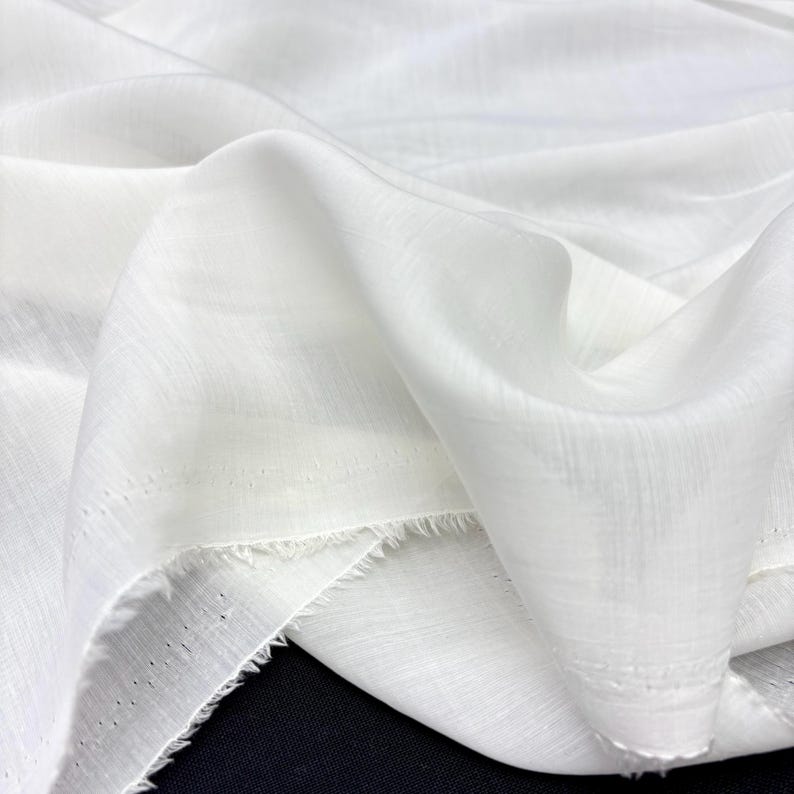 May include: Close-up of white fabric with a soft, flowing texture. The material is draped, showing folds and subtle variations in light and shadow. The edges of the fabric are unfinished, with a frayed appearance. The fabric appears to be lightweight and semi-sheer.