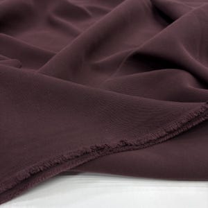 May include: A length of solid dark plum colored fabric with a visible twill weave texture. The material is draped in soft folds, showcasing a matte finish and a frayed, unfinished edge along the bottom corner of the textile piece.