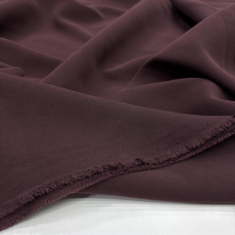 May include: A length of solid dark plum colored fabric with a visible twill weave texture. The material is draped in soft folds, showcasing a matte finish and a frayed, unfinished edge along the bottom corner of the textile piece.