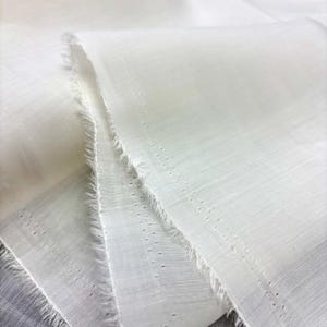 May include: Close-up of white fabric with a textured weave and frayed edges. The fabric appears lightweight and semi-sheer, with small, evenly spaced perforations. The folds and layers create shadows, highlighting the texture.