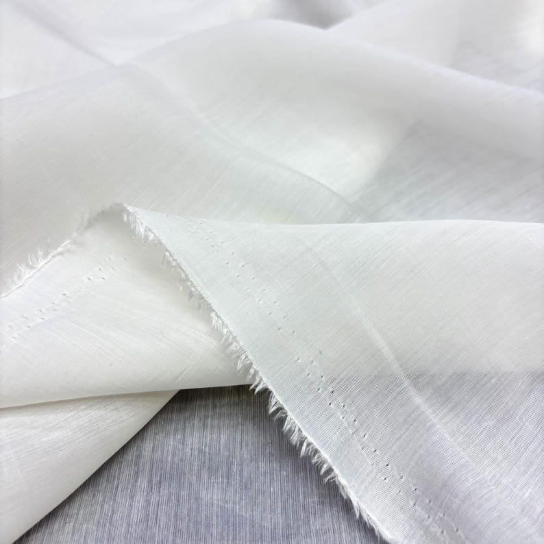 May include: Close-up of white fabric with a textured surface. The fabric is folded, showing the raw edge. The material appears lightweight and semi-sheer, suitable for clothing or home decor. The fabric is draped.