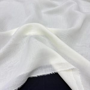 May include: Close-up of white fabric with a delicate woven texture. The fabric is draped, forming gentle folds and shadows. The edges are unfinished, with a frayed look. The material appears lightweight and semi-transparent, suitable for clothing.