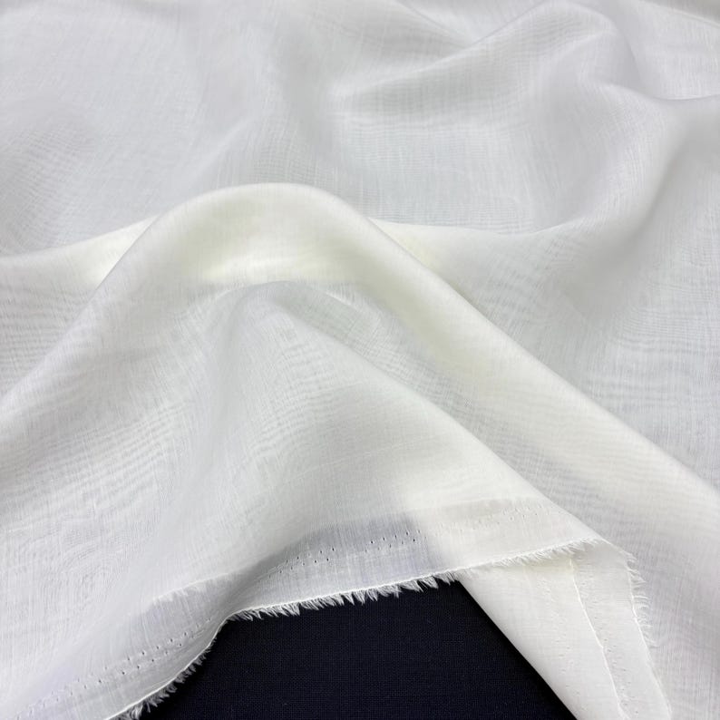 May include: Close-up of white fabric with a delicate woven texture. The fabric is draped, forming gentle folds and shadows. The edges are unfinished, with a frayed look. The material appears lightweight and semi-transparent, suitable for clothing.