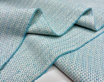 May include: A close-up shot of folded woven fabric. The fabric is a mix of light blue and white, with a textured weave. The edges are trimmed with a darker blue, and the weave pattern is clearly visible.