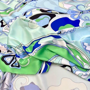 以下が含まれることがあります： 鮮やかな生地のクローズアップ。カラフルな抽象的なデザインです。生地は、水色、緑、白、黒の形が混ざっています。素材は絹のような光沢があり、高級テキスタイル、おそらくシルクまたはサテンを示唆しています。