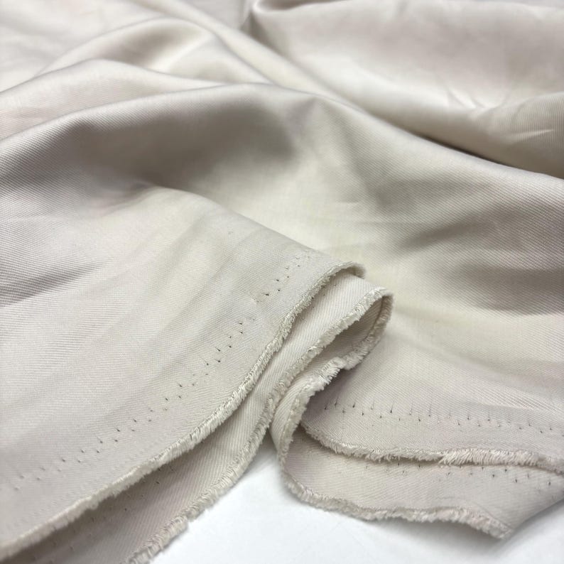 May include: A length of off-white fabric with a visible diagonal twill weave pattern. The material features a smooth, slightly lustrous surface with soft folds and drapes. The edges of the textile are unfinished and frayed, showing the raw weave.