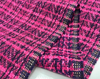 May include: Close-up of a vibrant pink and black woven fabric with a plaid pattern. The fabric has a textured appearance and is folded, revealing the edges with gold and black accents. Ideal for apparel and craft projects.