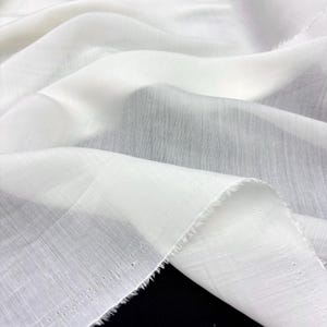 May include: Close-up of a flowing, white fabric with a delicate, semi-sheer appearance. The fabric is draped in soft folds, revealing a subtle texture and a raw, unfinished edge. The material appears lightweight and airy.