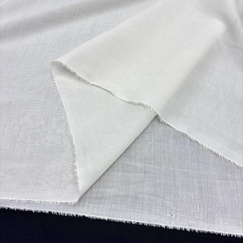 May include: Close-up of white fabric with a textured weave. The fabric is folded over, creating a layered effect. The edges are unfinished, with a frayed appearance. The fabric appears to be lightweight.