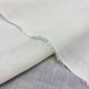 May include: Close-up of white fabric with a raw, frayed edge. The fabric appears lightweight and slightly translucent, with a visible woven texture. The material is folded over, creating a layered effect.