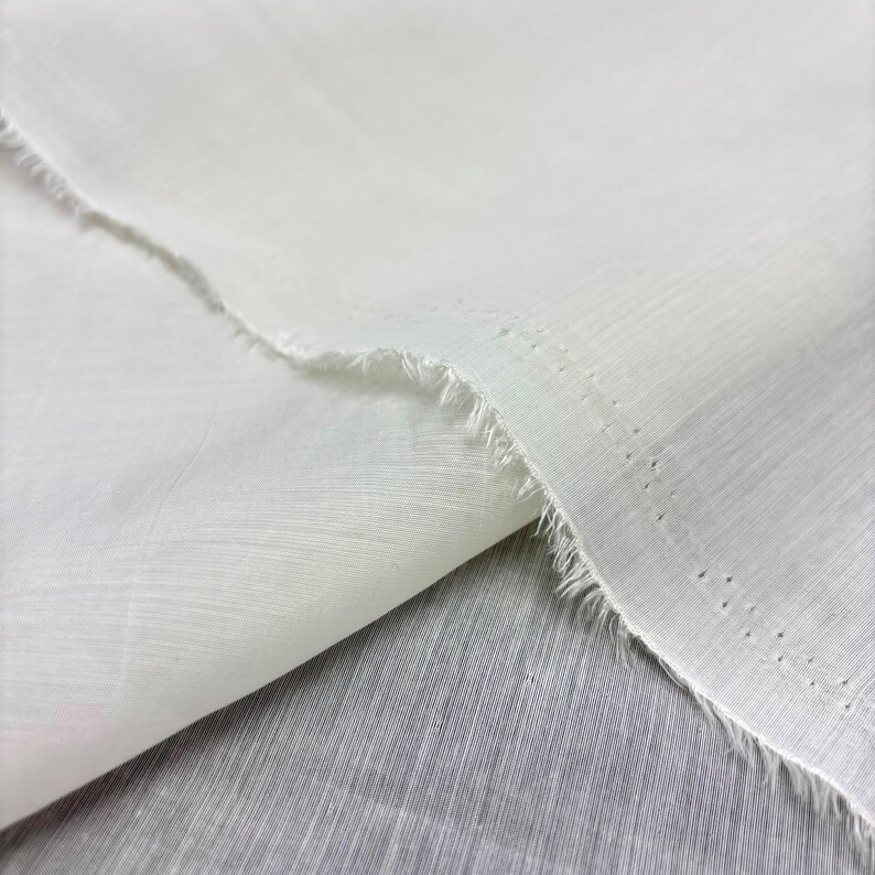 May include: Close-up of white fabric with a raw, frayed edge. The fabric appears lightweight and slightly translucent, with a visible woven texture. The material is folded over, creating a layered effect.