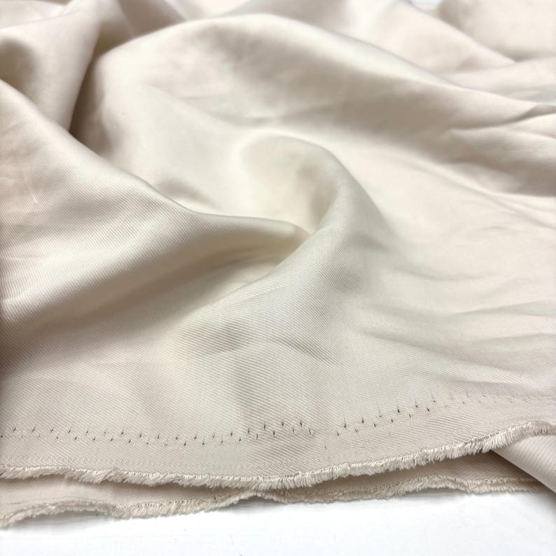 May include: A length of solid off-white fabric with a smooth, slightly lustrous finish and a visible diagonal twill weave pattern. The material features a raw, frayed edge along the bottom with a distinct row of small, dark, evenly spaced stitch marks.