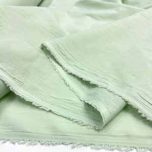 May include: A length of light mint green fabric with a soft, slightly textured surface and visible horizontal weave lines. The material features raw, frayed edges along the cut sides, showcasing the loose threads and the lightweight, semi-opaque quality.
