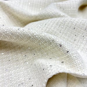 Puede incluir: Primer plano de una tela texturizada de color blanco roto con un brillo sutil. La tela está tejida con un tejido apretado y presenta pequeñas lentejuelas plateadas dispersas. El material está doblado, mostrando la textura y la caída de la tela.