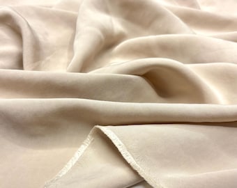 Puede incluir: Primer plano de una tela beige con pliegues suaves y un borde sin rematar. La tela tiene un aspecto liso y lujoso, lo que sugiere un material de alta calidad. El color es un tono claro y neutro.